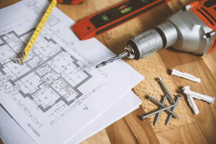 A floor plan on a wooden table next to a drill, screws and a tape measure
